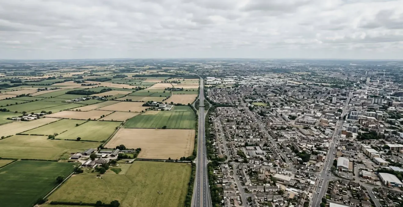 Vue aérienne minimaliste montrant la transition entre une zone rurale et une zone urbaine dense, symbolisant l'impact du changement d'adresse sur l'évaluation du risque d'assurance
